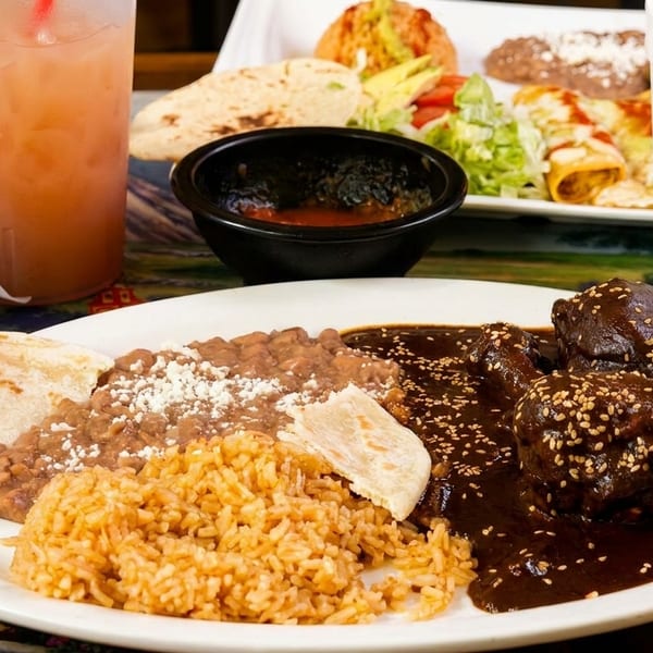 Plates of Mexican food, including mole poblano, rice, beans, and enchiladas, with drinks on a colorful table.