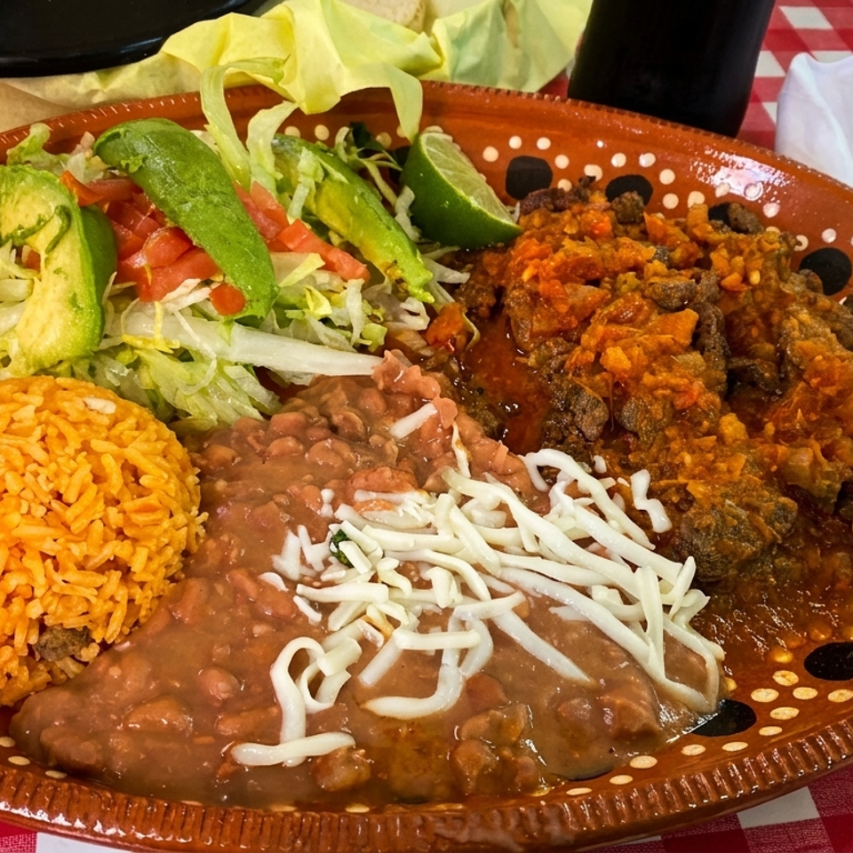A Mexican dish with rice, beans, shredded cheese, salad, and meat in red sauce on a brown plate.