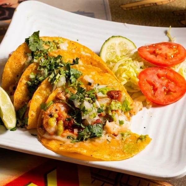 Three shrimp tacos with cilantro, onions, and avocado, served with lime, lettuce, and tomato.