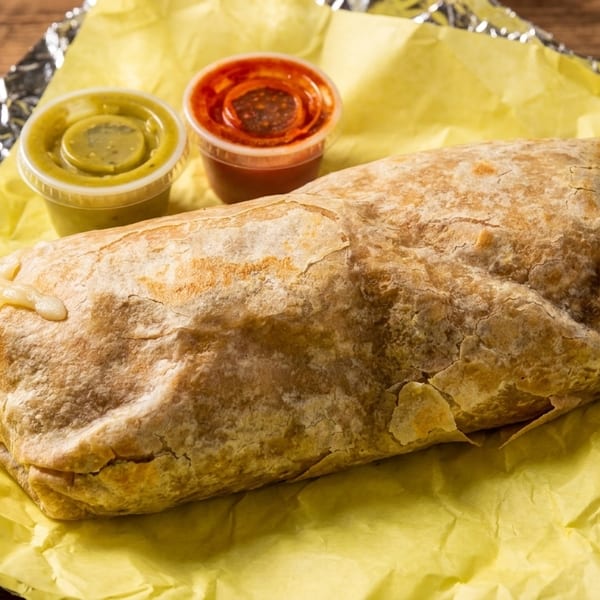 Large burrito wrapped in yellow paper and foil, with two small containers of salsa on a wooden table.